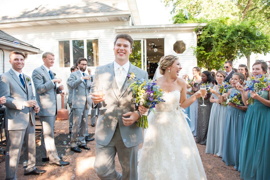 Bride and groom exit the farm kitchen while guest blow bubbles